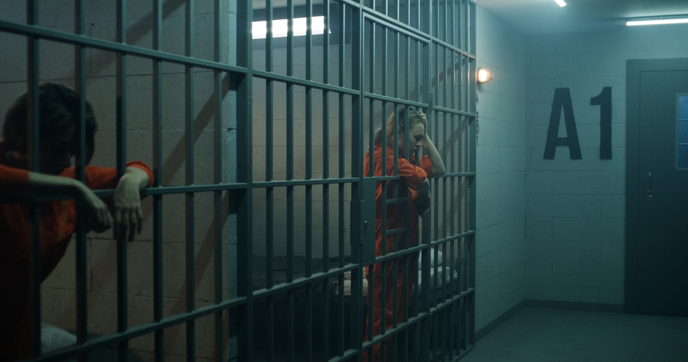 One,Female,Prisoner,In,Orange,Uniform,Stands,Behind,Metal,Bars,
